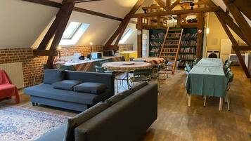 Una habitación estilo loft con techo alto, vigas de madera a la vista, una mesa redonda con sillas, un sofá, una estantería y un radiador.