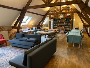 A loft-style room with a high ceiling, exposed wooden beams, a round table with chairs, a sofa, a bookshelf, and a radiator.