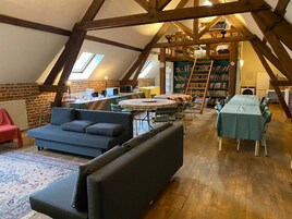 Una habitación estilo loft con techo alto, vigas de madera a la vista, una mesa redonda con sillas, un sofá, una estantería y un radiador.