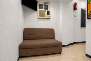 Un sofá pequeño color marrón en un pasillo con paredes blancas, piso de baldosas, una ventana con persianas y un extintor de incendios colgado en la pared.