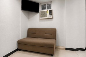 Un sofá pequeño y marrón en una esquina de una habitación con paredes blancas, un aire acondicionado instalado y un televisor de pantalla plana en la pared.