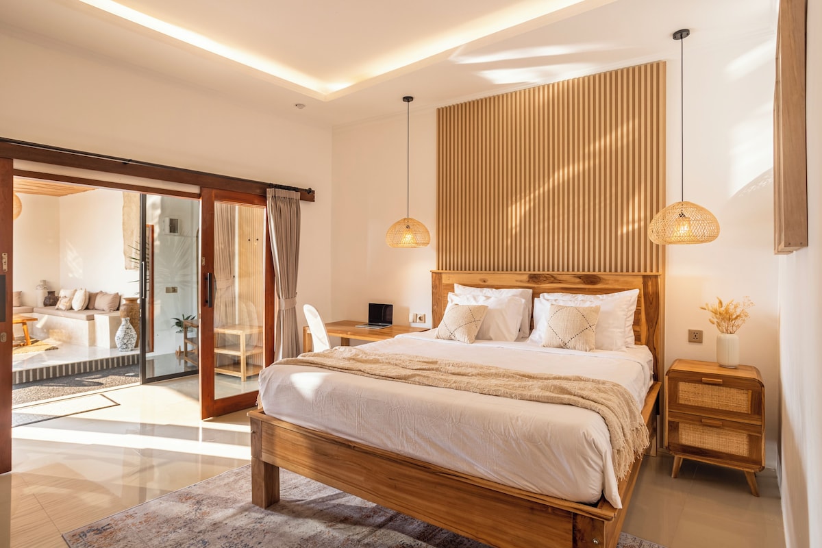 A modern bedroom with a large bed, wooden headboard, two bedside tables, and a sliding glass door leading to a balcony.