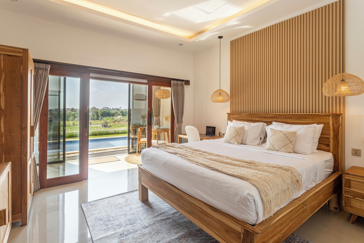 A bedroom with a large bed, a wooden headboard, a view of a pool, and a dining area with a table and chairs.