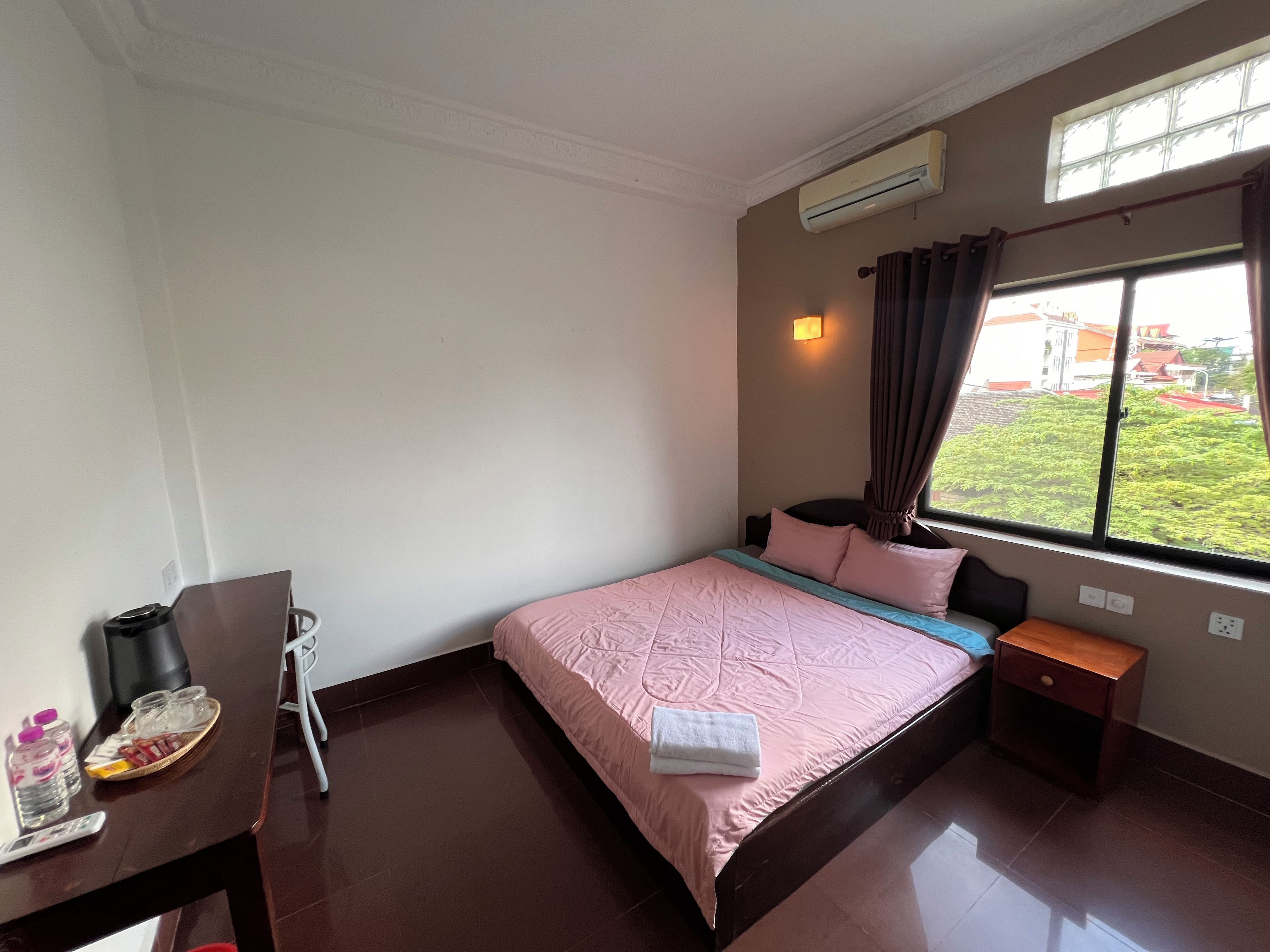 A hotel room with a bed, bedside table, a desk with a chair, an air conditioner, and a window with a view of greenery.