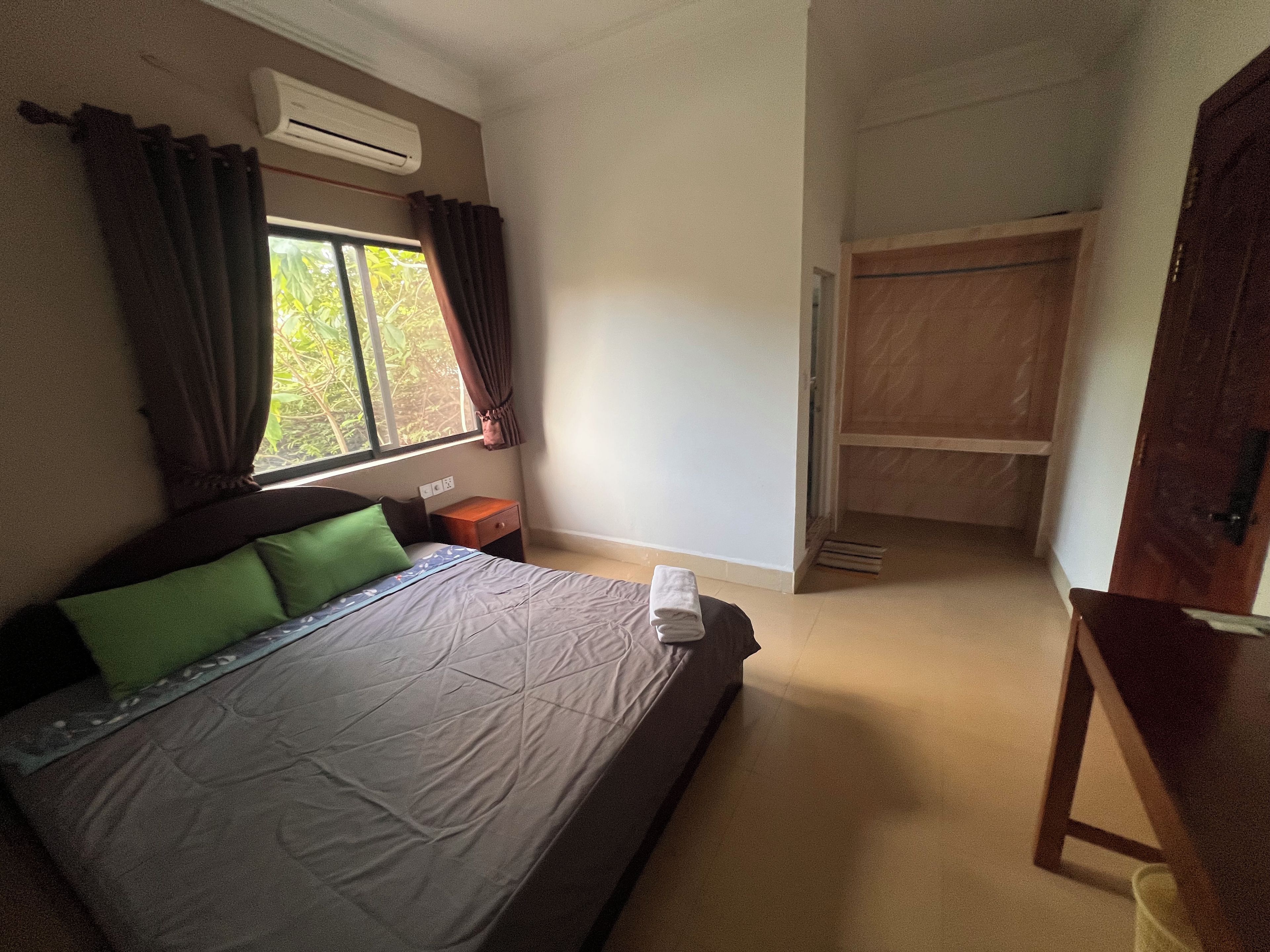 Un dormitorio con cama, mesita de noche, equipo de aire acondicionado, ventana con cortinas y armario de madera.