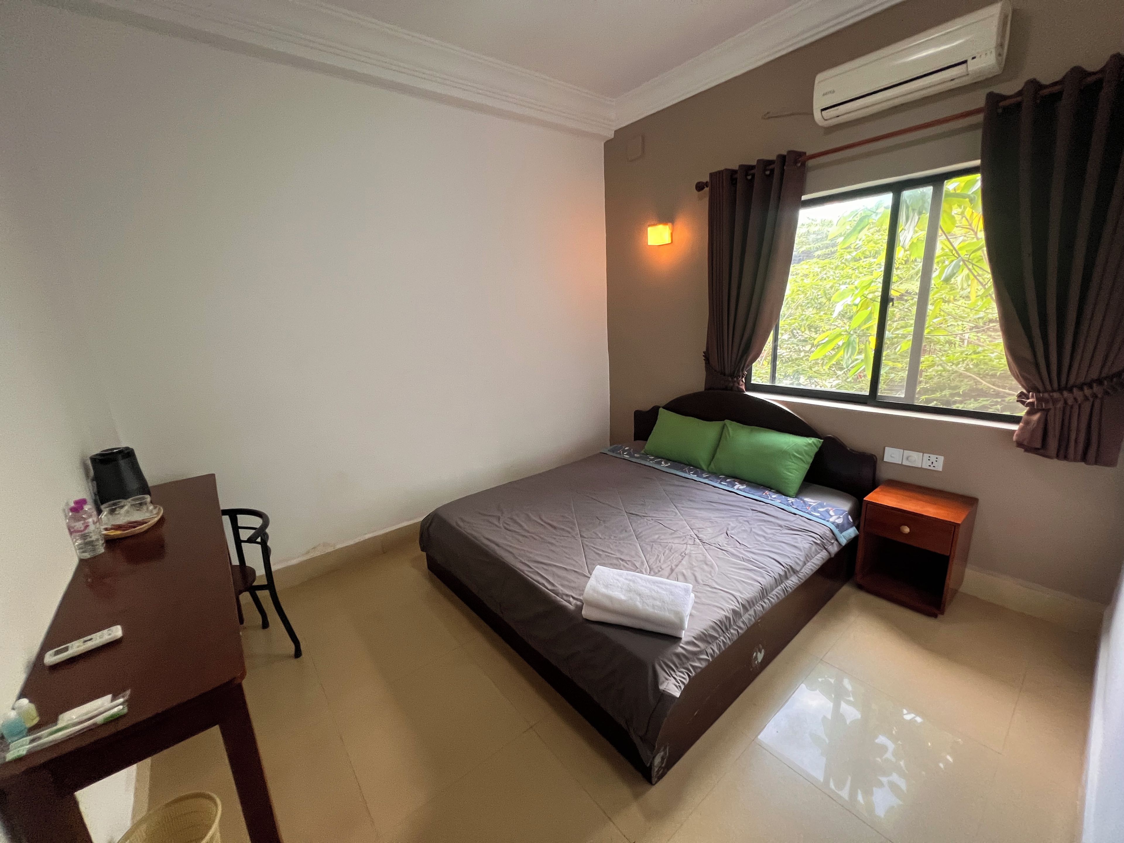 A bedroom with a bed, a bedside table, a desk, a chair, an air conditioning unit, and a window with curtains.
