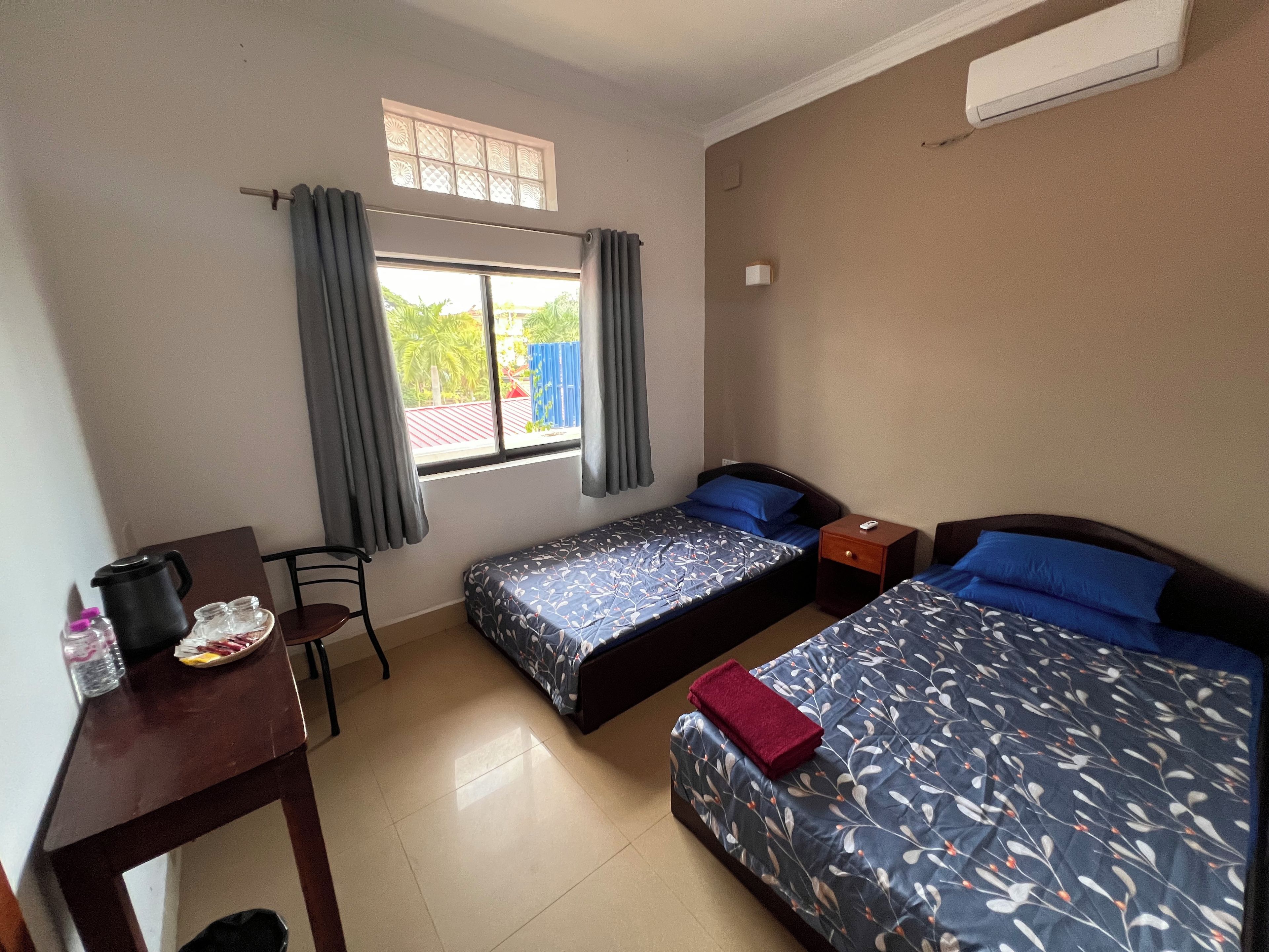 Habitación de hotel con dos camas individuales, un escritorio con silla, una mesita con tetera y una ventana con cortinas.