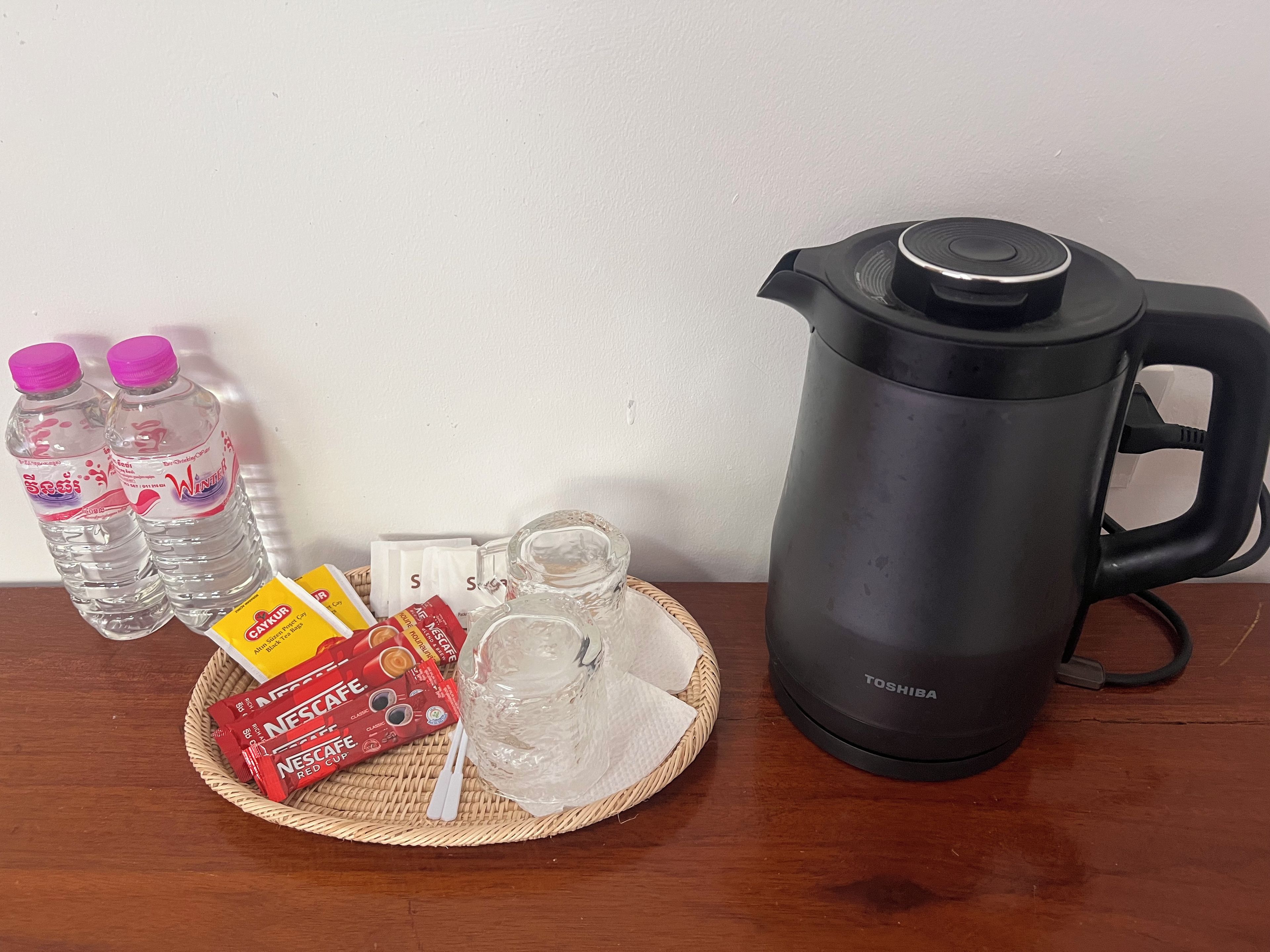 Uma mesa com uma chaleira elétrica preta, duas garrafas de água, um jarro de vidro, um tapete tecido e alguns sachês de creme para café.