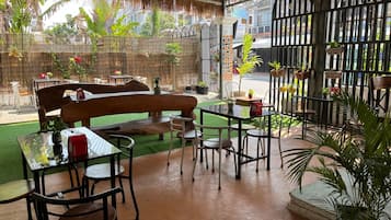 An outdoor seating area with tables, chairs, and a bench, surrounded by potted plants and a bamboo fence.