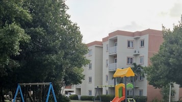Een speeltuin met schommels en een glijbaan, omringd door gras en bomen, met een gebouw op de achtergrond.
