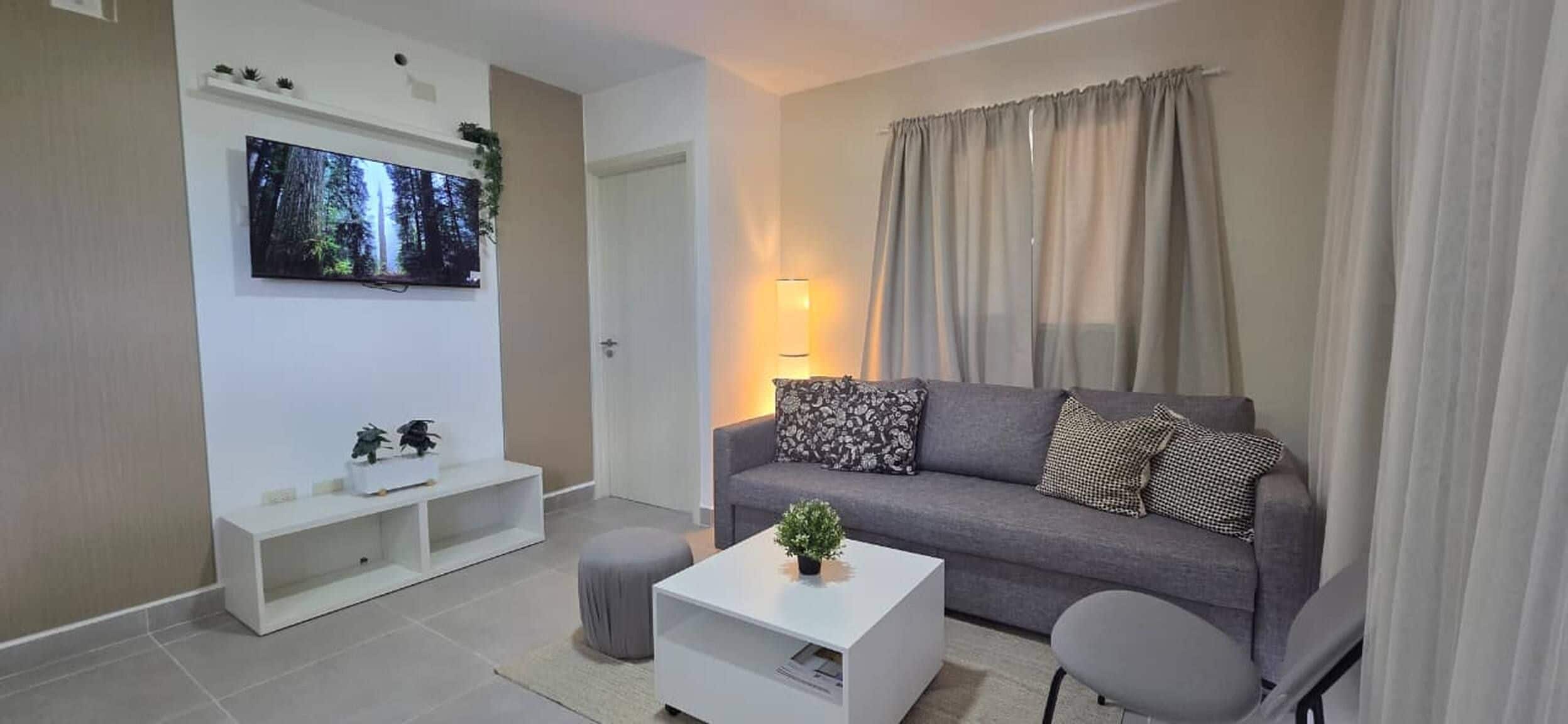 Een woonkamer met een grijze bank, een witte salontafel en een flatscreen televisie aan de muur.