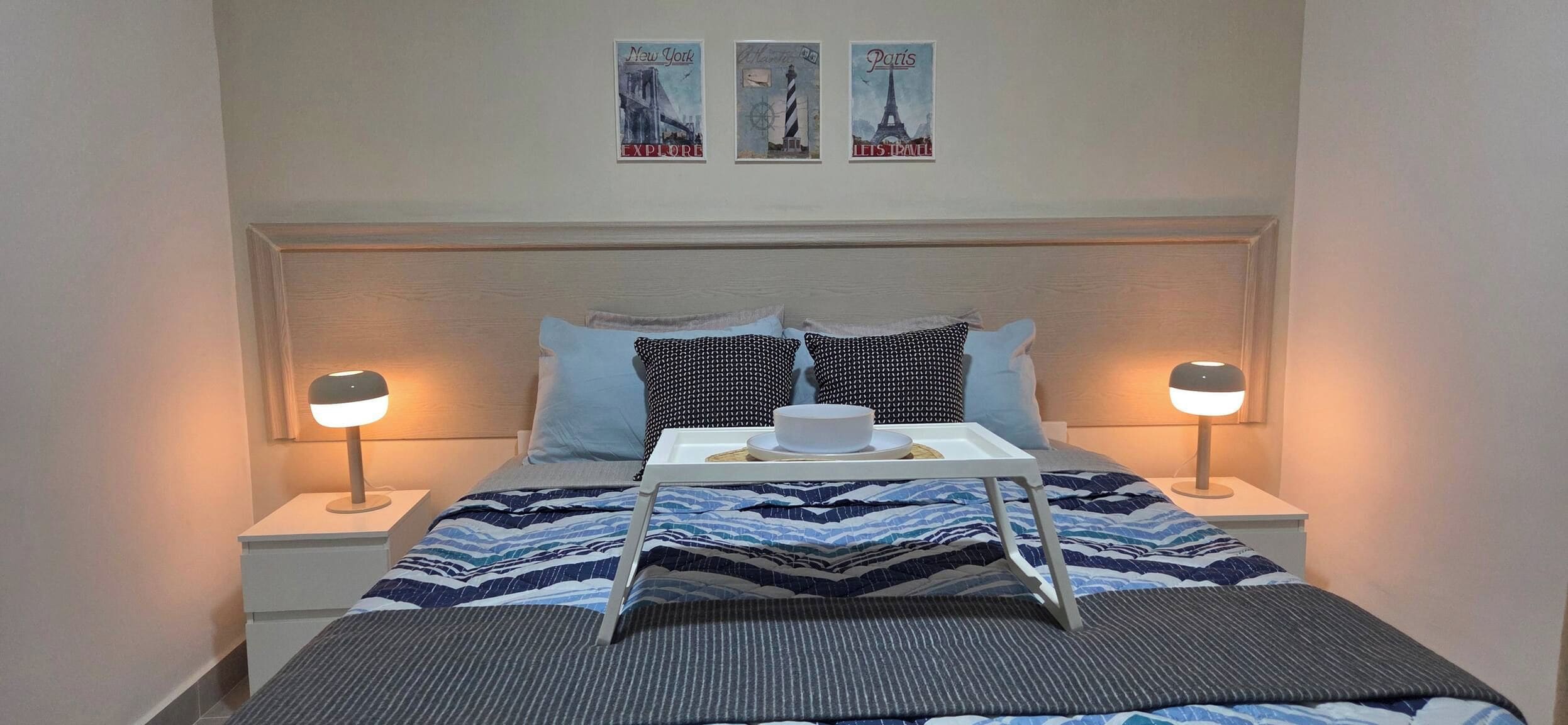 Een netjes opgemaakt bed met een tray ontbijtartikelen, twee bedlampjes en drie posters met reisthema aan de muur.