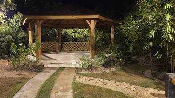 A wooden gazebo with a concrete pathway leading to it, surrounded by greenery.