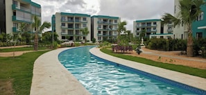 A swimming pool with a curved edge, surrounded by a well-maintained lawn and palm trees. - Serena Retreat in Punta Cana Nature View Apartment With 11 Pools (Punta Cana)