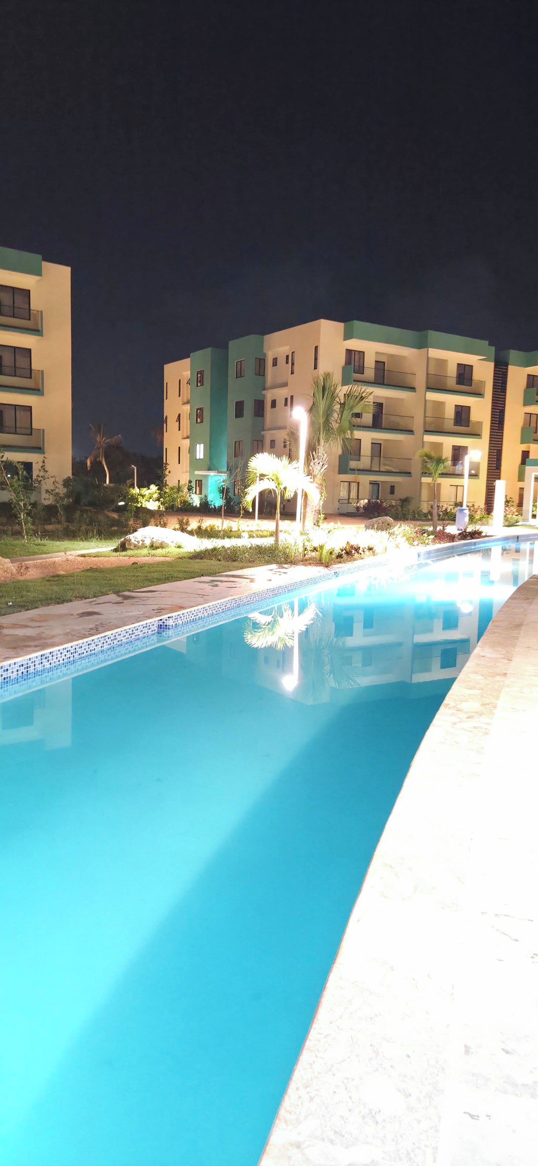 Una piscina con superficie de agua azul cristalina, rodeada por un jardín bien cuidado con palmeras y arbustos.