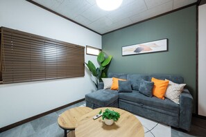Quarto com sofá em formato de L, mesa redonda de madeira, planta em vaso e uma janela grande com persianas.