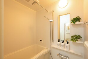 A bathroom with a bathtub, a shower, a mirror, and shelves with toiletries. - Zeroichi Hotel House Hakata (Fukuoka)