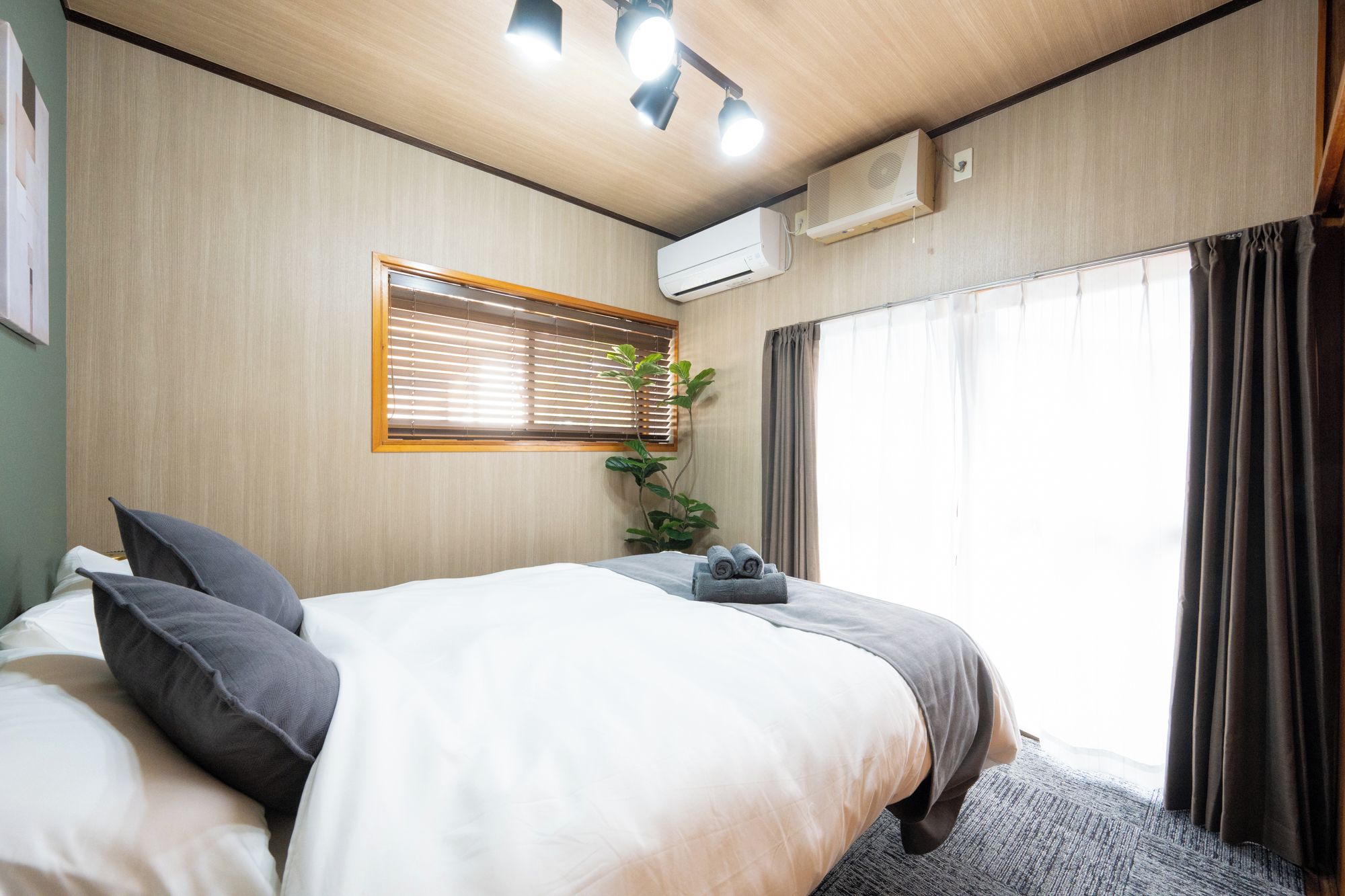 A hotel room with a bed, a window with blinds, an air conditioning unit, and a plant.