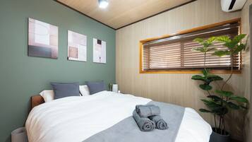 A hotel room with a bed, a window with blinds, a potted plant, and artwork on the wall.