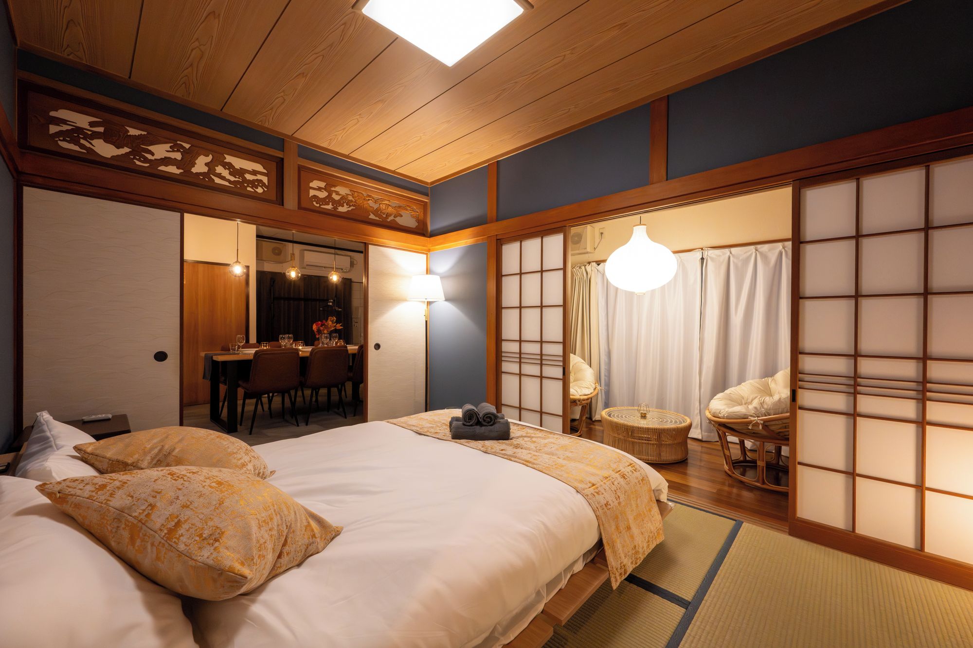 A traditional Japanese-style room with tatami flooring, a bed with a white and yellow bedspread, a dining area with a table and chairs, and a sliding shoji screen.
