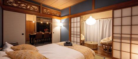 Una habitación tradicional japonesa con suelo de tatami, una cama con colcha blanca y amarilla, un rincón de comedor con mesa y sillas, y una pantalla corrediza shoji.