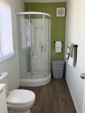 A bathroom with a glass-enclosed shower, a toilet, a trash bin, and a towel rack with towels.