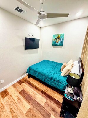 Een slaapkamer met een bed, een plafondventilator, een televisie, een schilderij en een nachtkastje met een lamp.