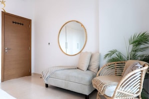 A bedroom with a wooden door, a round mirror, a bed with striped bedding, a wicker chair, and a potted plant. - 2BR | Balcony | Yas Island | Pool & Gym (Abu Dhabi)