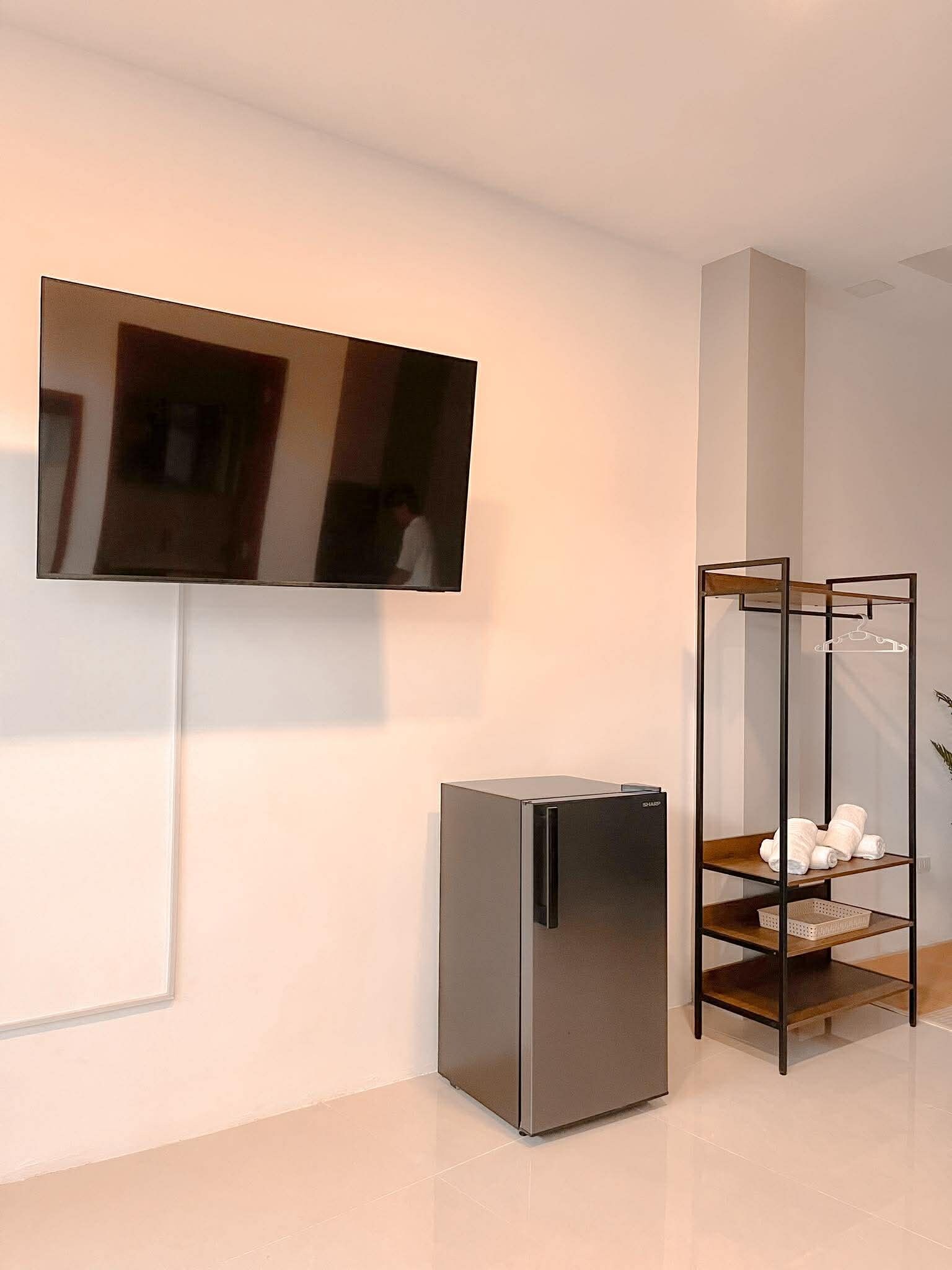 Una habitación moderna con un televisor de pantalla plana montado en la pared, un refrigerador empotrado y un estante de madera con toallas.