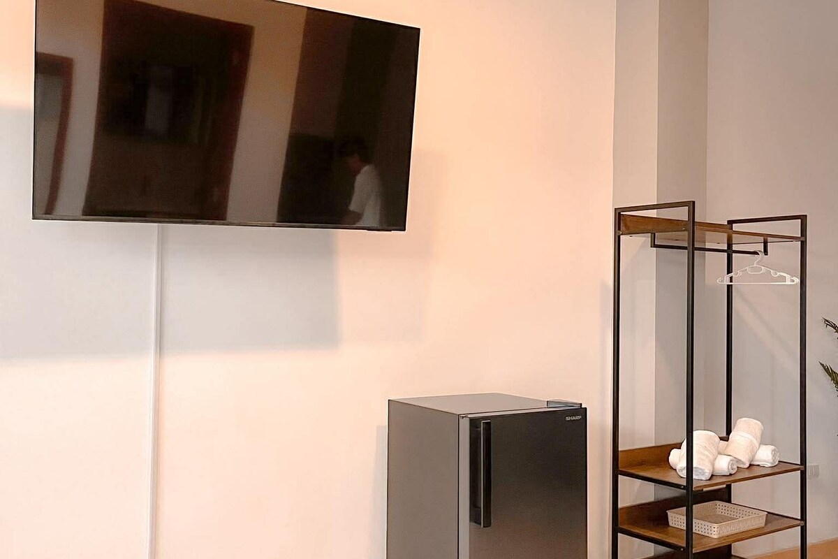 A modern room with a flat-screen TV mounted on the wall, a built-in refrigerator, and a wooden shelf unit with towels.