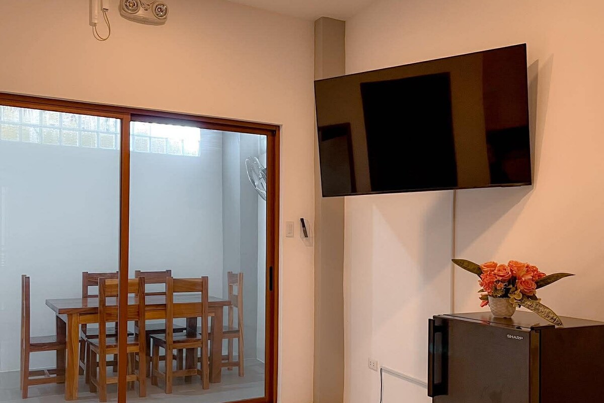 A modern kitchen with a refrigerator, a flat-screen TV mounted on the wall, and a dining area with wooden chairs and a glass door leading to a patio.