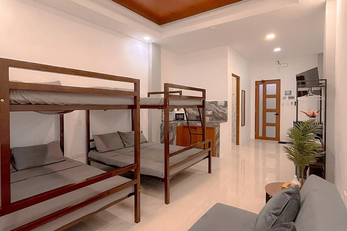 A modern hotel room with bunk beds, a sofa, a small kitchen area, and a hallway leading to other rooms.