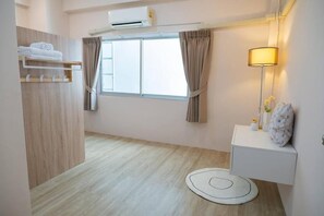 A hotel room with a wooden floor, a window with curtains, a white bedside table with a lamp, and a towel rack. - Lumi Minimal Residence (Krung Thep Maha Nakhon)