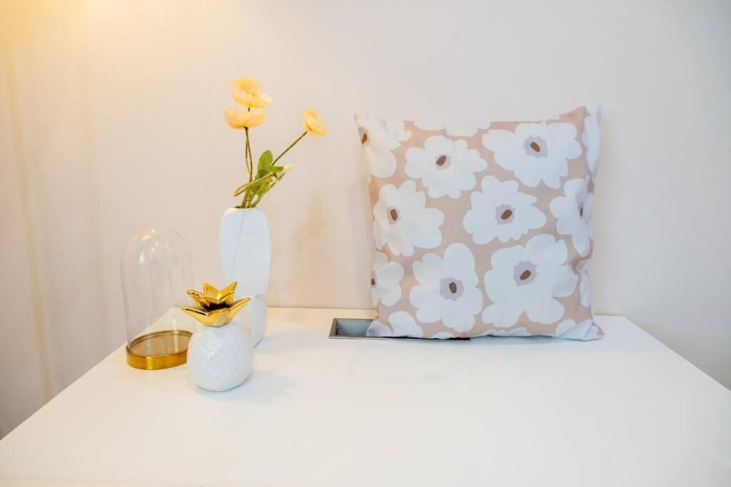 Una mesita de noche blanca con un jarrón de flores amarillas, una campana de cristal transparente y un jarrón blanco con un adorno dorado.
