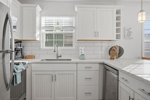 A modern kitchen with white cabinetry, a stainless steel refrigerator, a sink, and a window with blinds. - Garnele's retreat- come relax (Galveston)