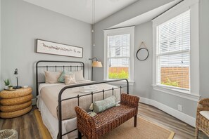 Une chambre à coucher avec un lit, un banc, une lampe, un tableau au mur et une fenêtre avec des stores.