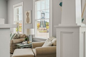 A living room with two sofas, a table, and a lamp. There are windows with blinds, a wall-mounted artwork, and a view of a building outside. - Old Flamingo Beach House (Galveston)