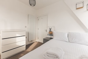 A bedroom with a bed, a dresser, a radiator, a door, and a small table with a lamp and decorative items. - Bethnal Green Penthouse & Terrace (London)