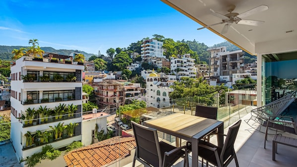 A balcony with a table and chairs, overlooking a cityscape with various buildings and greenery.