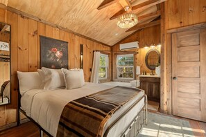 A wooden cabin bedroom with a large bed, a chandelier, a painting, and a bathroom with a mirror and sink. - Creek Mornings, Fire-Pit Nights • Cozy Cottage Near Thunder Ridge (Blue Eye)