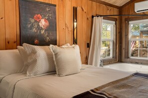 Interior de cabana de madeira, com cama, pintura, janela com cortinas e banheira.