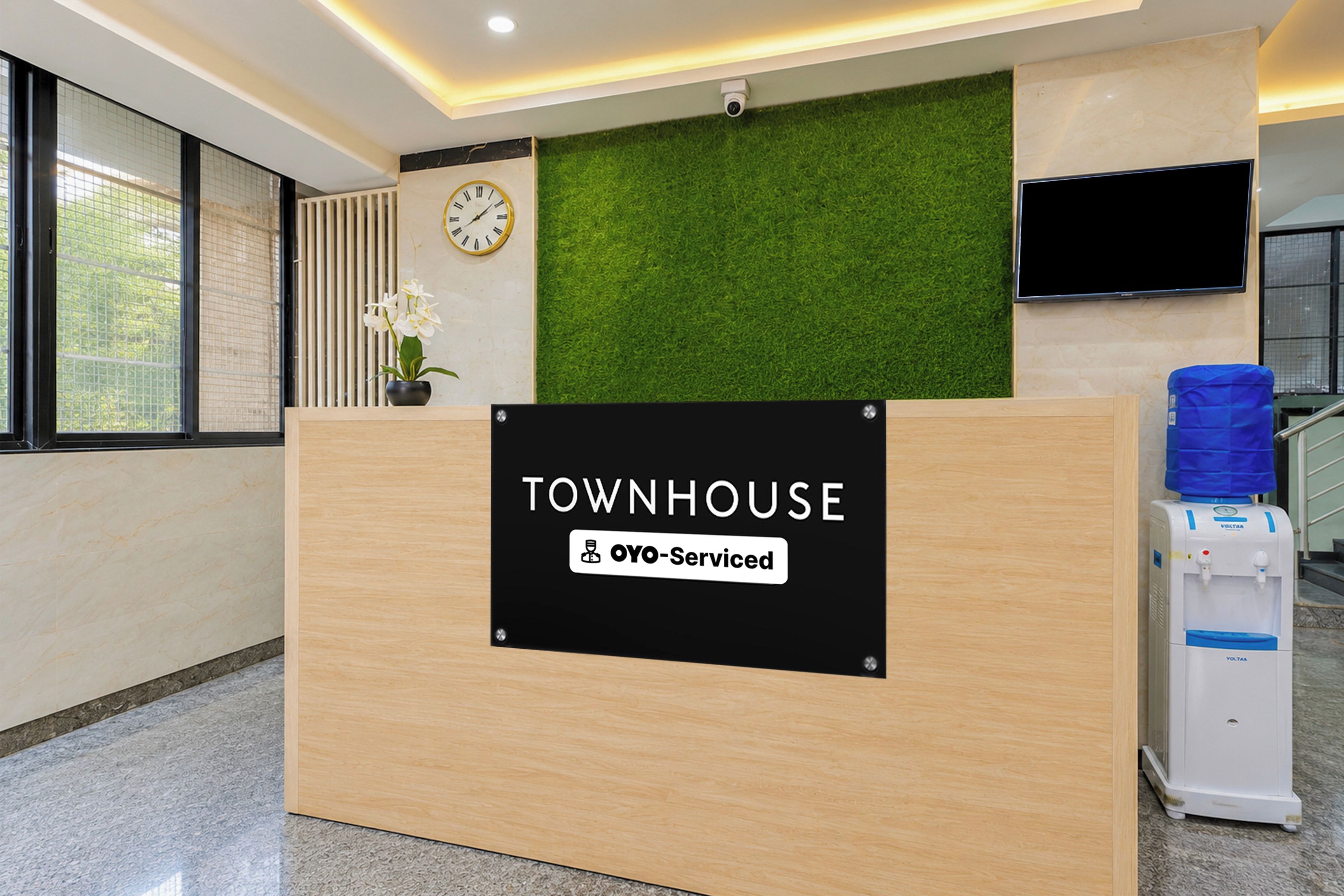 En receptionsdisk med en skylt som visar texten ”TOWNHOUSE OYO-Serviced” i en modern lobby med en grön vägg, en klocka och en vattenautomat.
