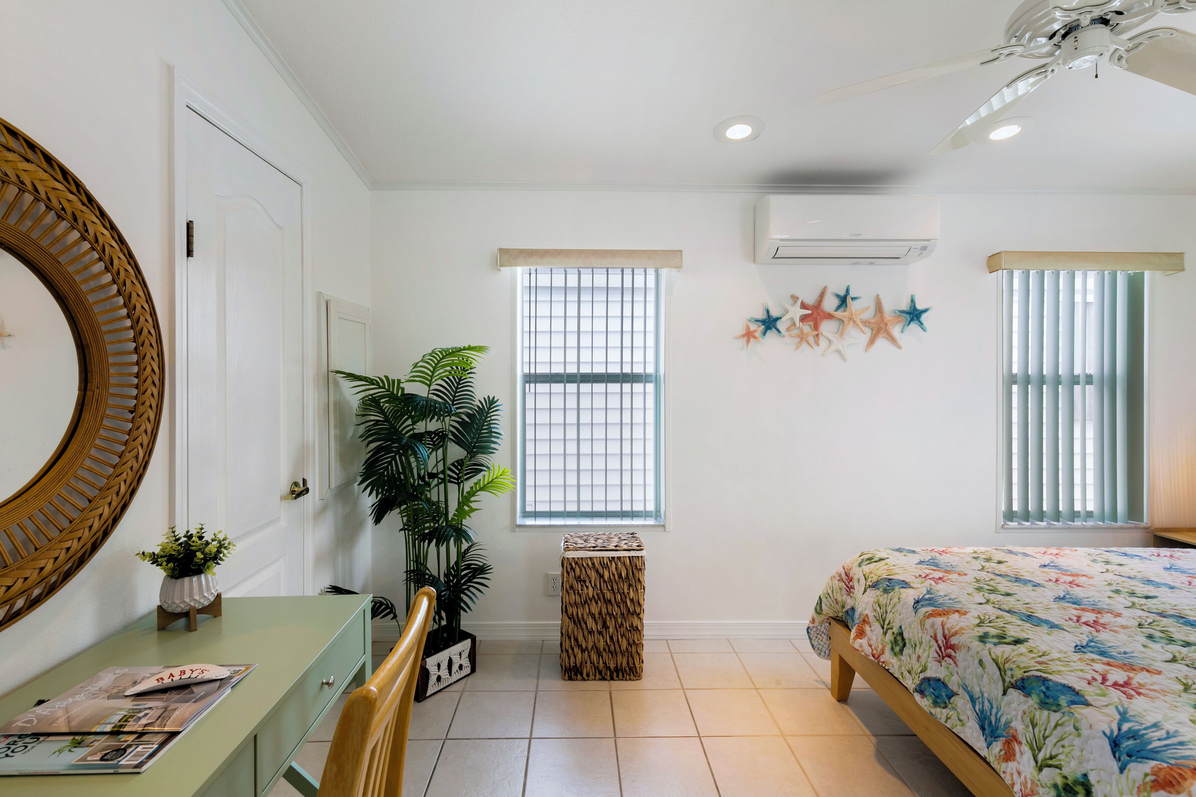 A bedroom with a bed, a desk, a chair, a potted plant, a mirror, and a window with blinds.