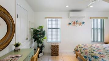 A bedroom with a bed, a desk, a chair, a potted plant, a mirror, and a window with blinds.