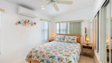 A bedroom with a bed, a ceiling fan, an air conditioning unit, a window with blinds, and a nightstand with a lamp.