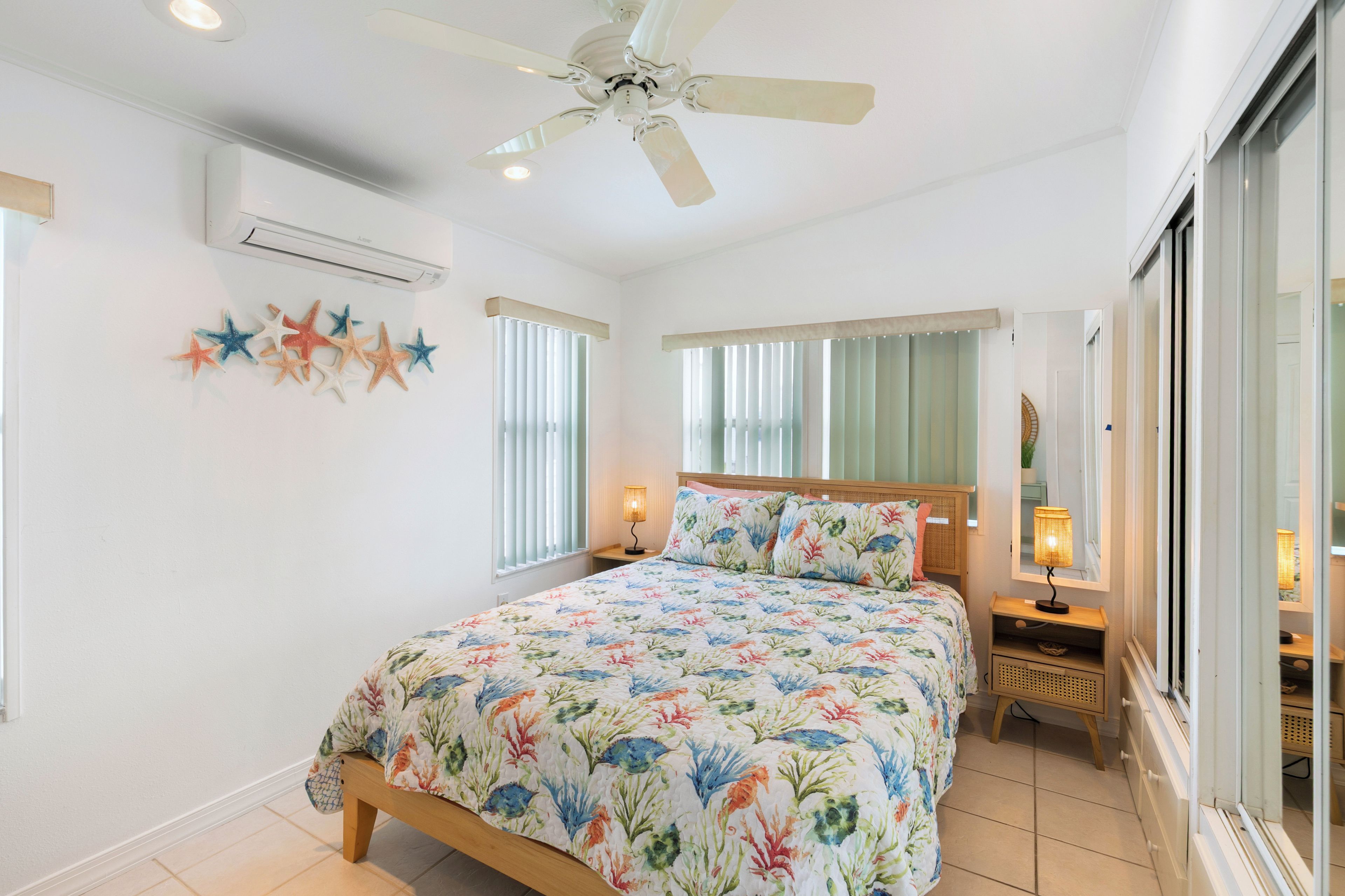 A bedroom with a bed, a ceiling fan, an air conditioning unit, a window with blinds, a bedside lamp, and a nightstand.