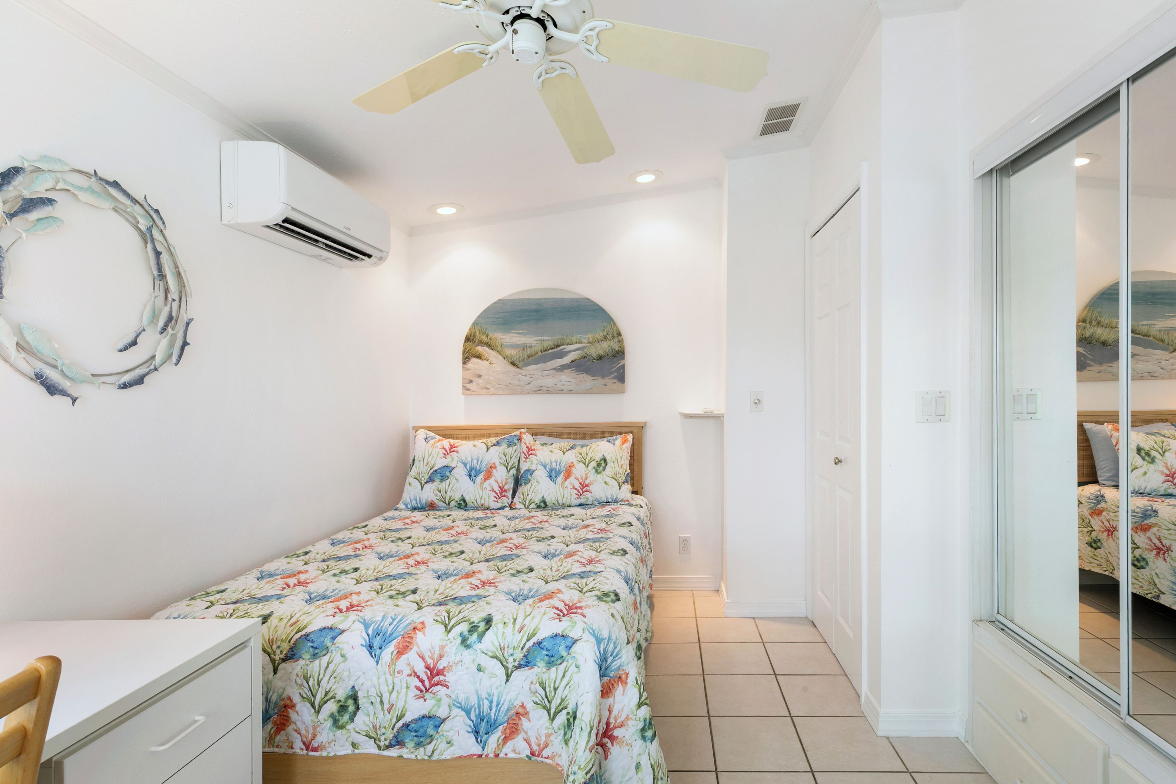 Een slaapkamer met een bed, bureau, stoel, plafondventilator, airconditioning en een schilderij met een strandthema aan de muur.