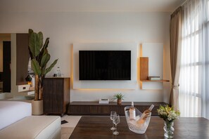 A modern living room with a flat-screen TV mounted on the wall, a wooden cabinet, a sofa, a coffee table with a bottle of champagne and glasses, and a potted plant. - 2 BR Apartement Near Thomas Beach, Uluwatu (South Kuta)