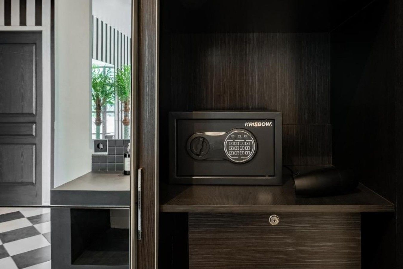 A secure safe with a digital keypad, placed on a wooden cabinet in a room with a checkered floor and a view of a balcony with plants.
