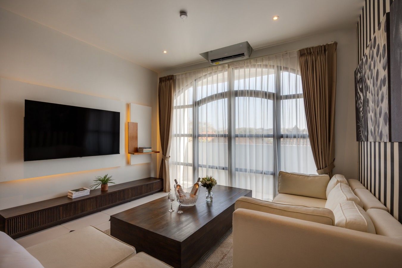 A modern living room with a flat-screen TV, a sofa set, a coffee table with decorative items, and a large window with curtains.
